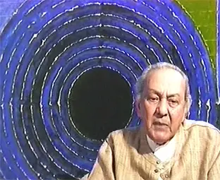 S.H. Raza, an elderly man with white hair, sits in front of one of his paintings, a dark circle at the centre of blue rings.