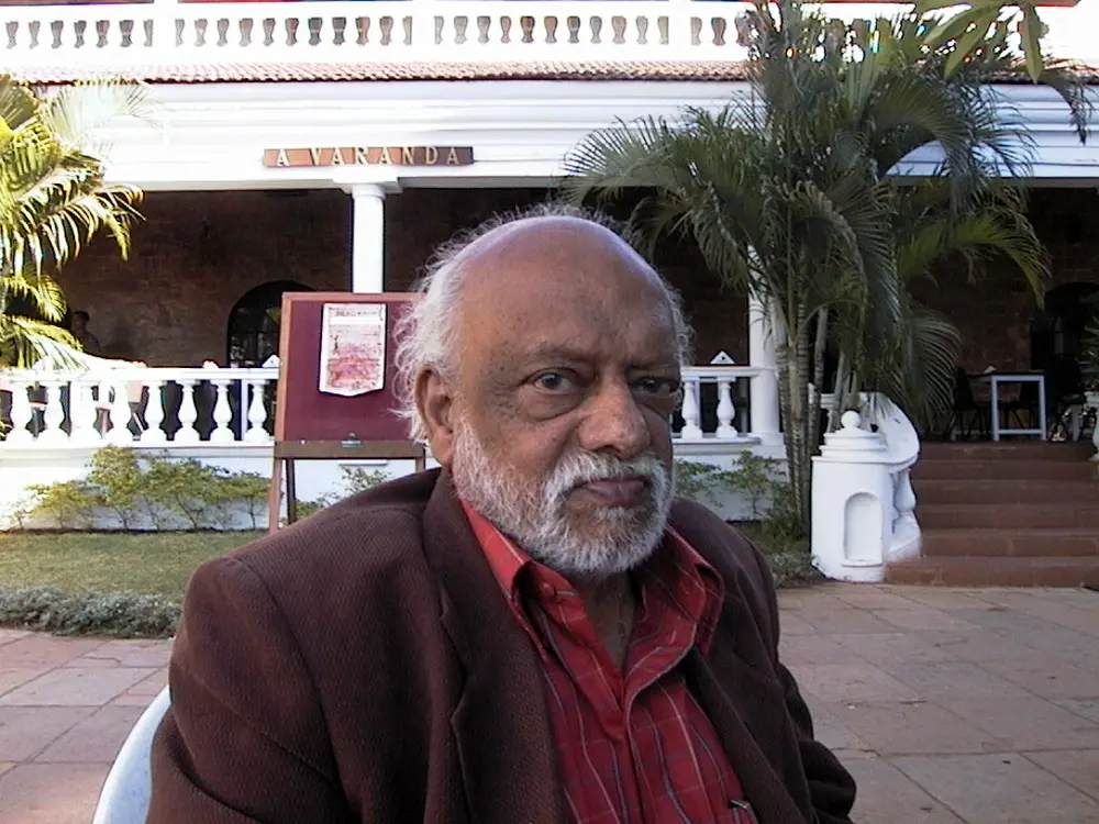 An elderly F.N. Souza, bald with a white beard, wearing a red checked shirt and brown jacket, sitting outside a hotel in Goa.