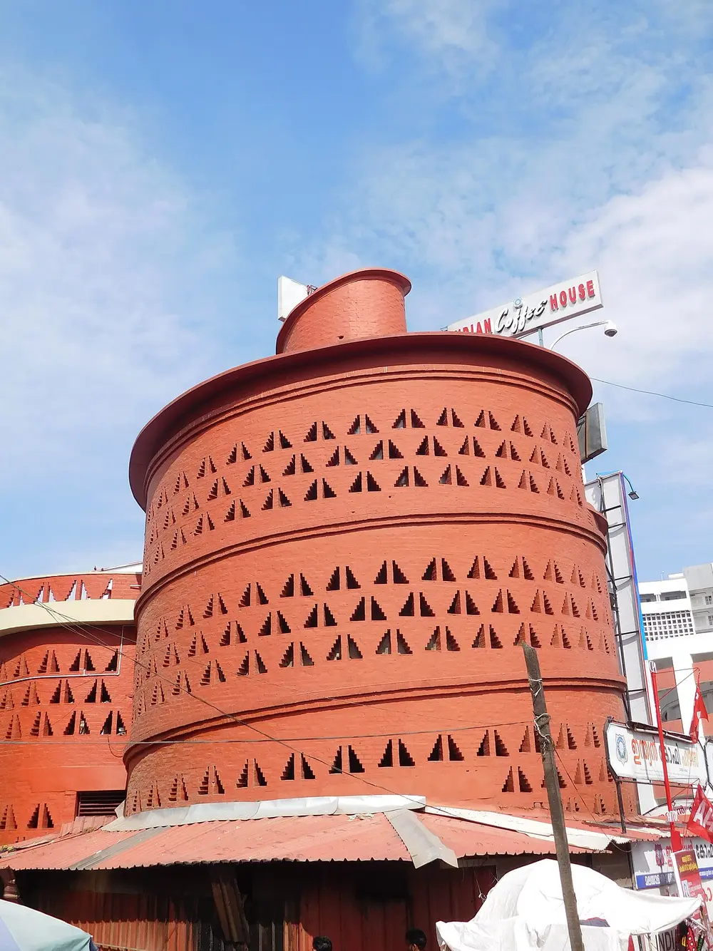A round tower of bare red brick with rows of small triangular holes in its walls, a wide rim around its top and a smaller cylinder on the roof, next to a sign reading Indian Coffee House.