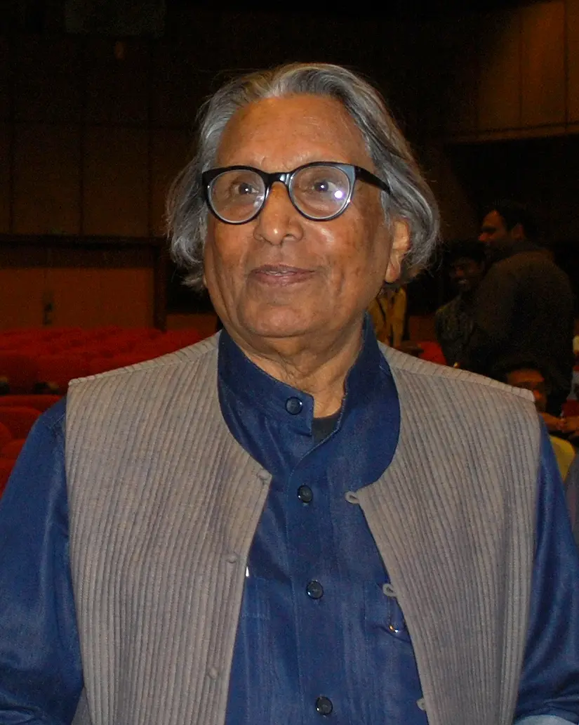 A portrait of B.V. Doshi, an elderly man with swept-back grey hair and round black glasses, wearing a blue kurta and a grey sleeveless jacket.