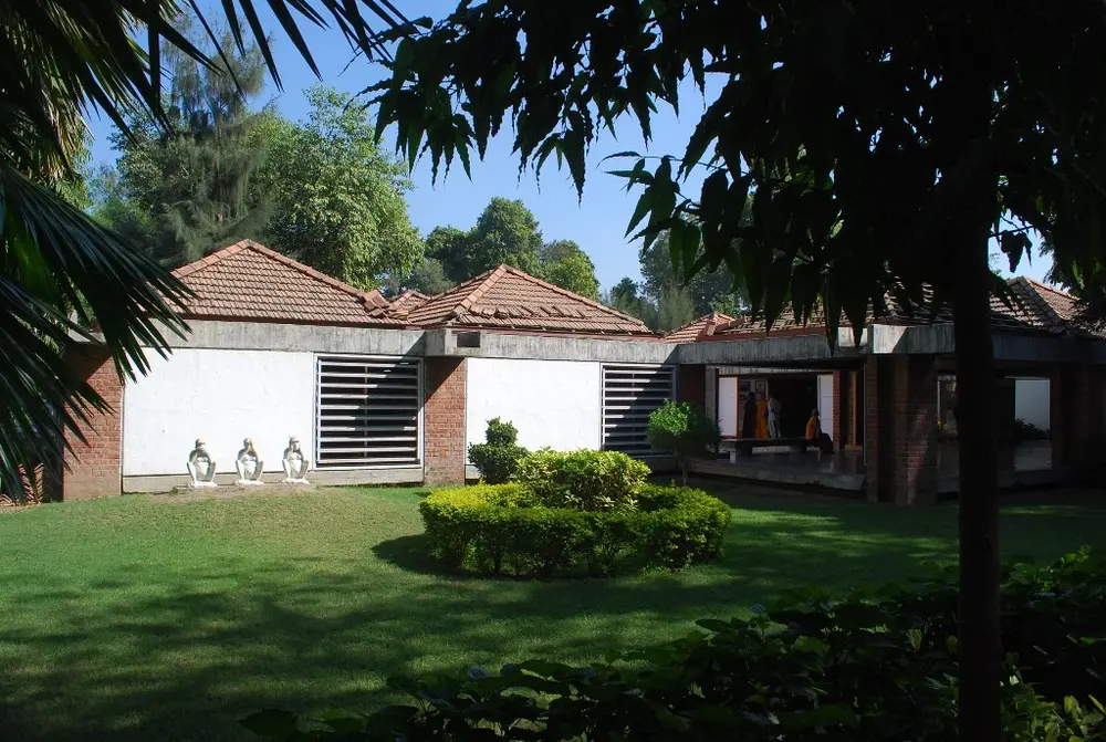 A low museum building with red tiled pyramid roofs resting on brick pillars, white wall panels and wooden louvred windows without glass, behind a green lawn with three small white statues of the three wise monkeys.