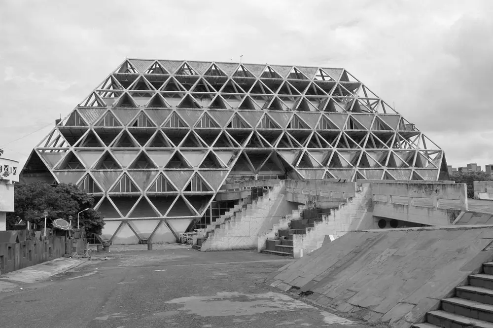 A black and white photo of a huge truncated pyramid made of a lattice of concrete triangles, with ramps and steps leading up to it across an empty concrete plaza.
