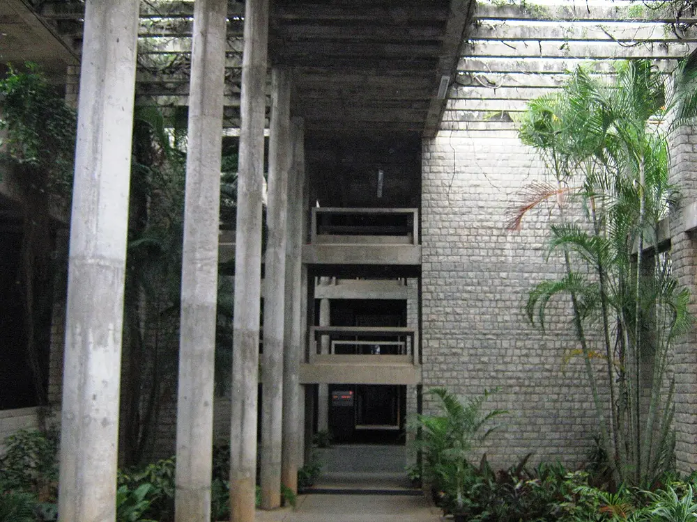 A tall, shady corridor at IIM Bangalore with a row of slim concrete columns on the left, a rough grey stone wall on the right, palm plants, and an open concrete pergola letting light in from above.