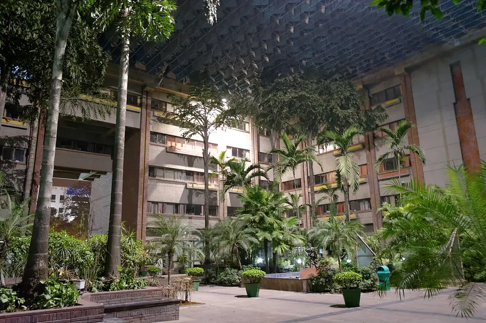 A courtyard at the India Habitat Centre at night, full of palm trees and plants, with brick and concrete office blocks rising on every side and a lattice of shading panels stretched high overhead.