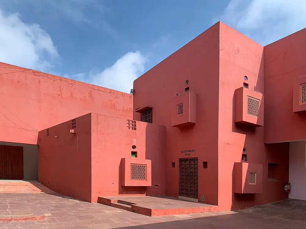 Red plastered walls of the Jawahar Kala Kendra around a paved courtyard, with small box-shaped windows covered by stone lattice screens, carved wooden doors and a blue sky.