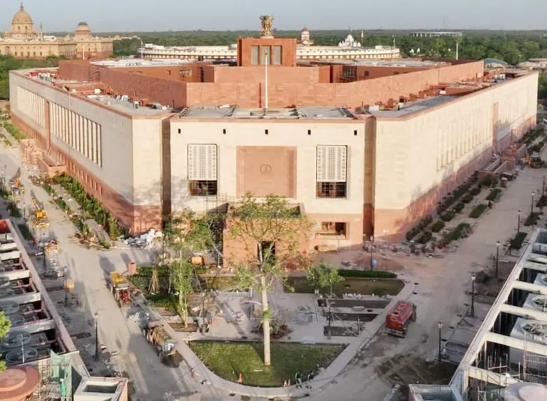 An aerial view of the new Parliament building in New Delhi, a large triangular building in red and cream sandstone, with landscaping still being finished around it.
