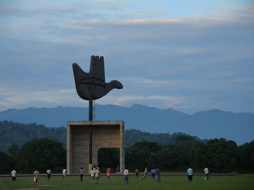 A giant dark metal sculpture of an open hand, shaped a little like a bird, standing on a tall pole above a concrete frame, with green hills behind and people playing football on the lawn in front.