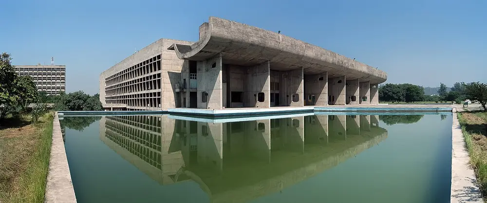 A huge grey concrete building reflected in a long pool, with a deep curved roof like an upturned umbrella resting on a row of thin concrete walls, and a grid of sun-shading boxes on its side.