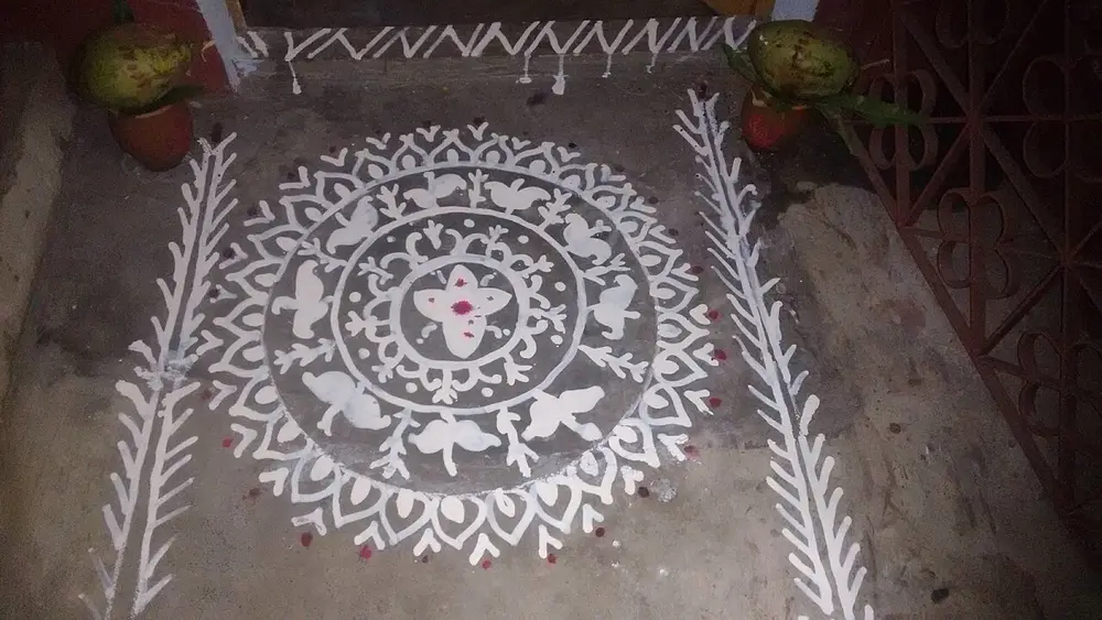 A white design painted on a grey doorstep at night: a round medallion of rings, petals and small birds, between two long leafy borders, with a pot topped by a green coconut and mango leaves at each corner.