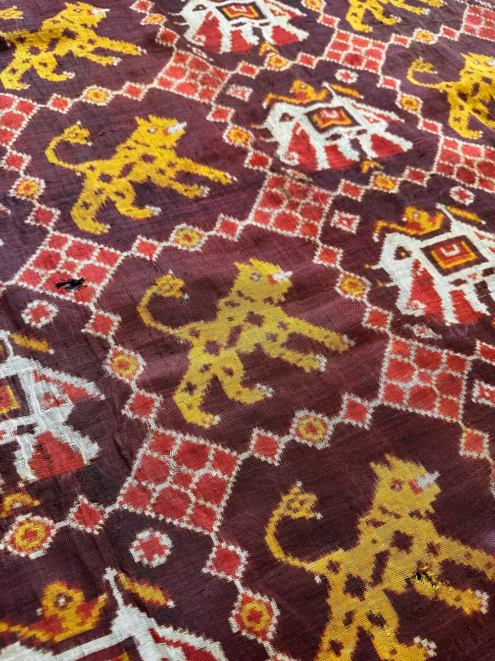 A close view of a silk cloth in deep red, with rows of white elephants carrying decorated saddle cloths and yellow tigers, all with soft, stepped edges, between small diamond patterns.