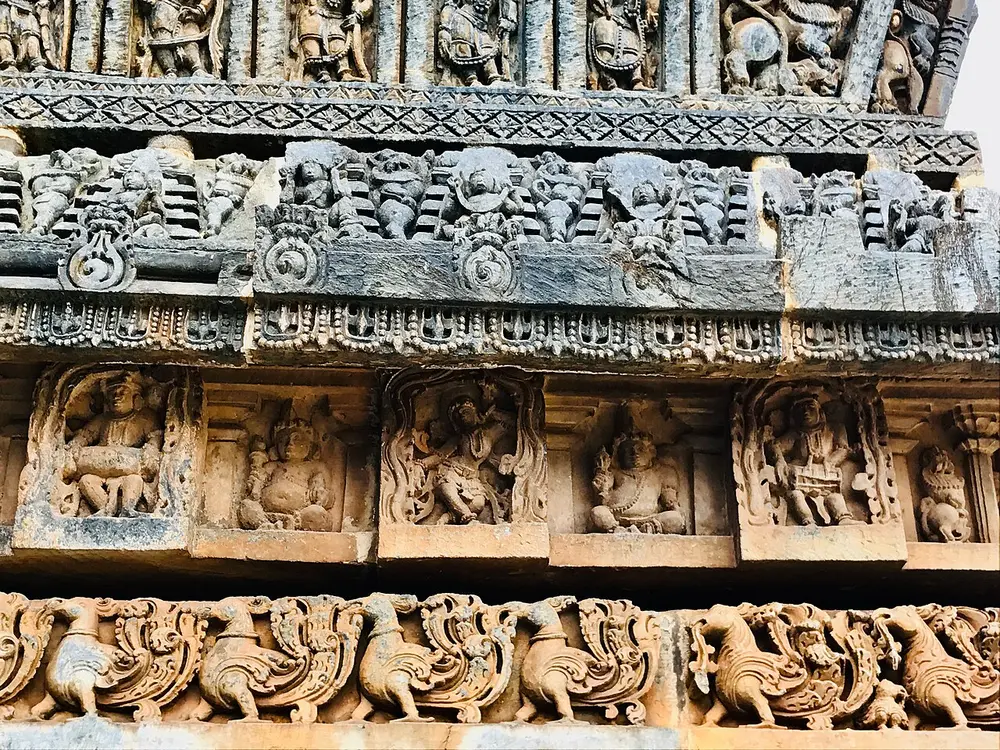 Bands of grey stone carving on a temple wall. At the bottom runs a row of plump birds with curling tails; above it, a row of small dancers and musicians in panels; higher up, figures and pierced stonework.