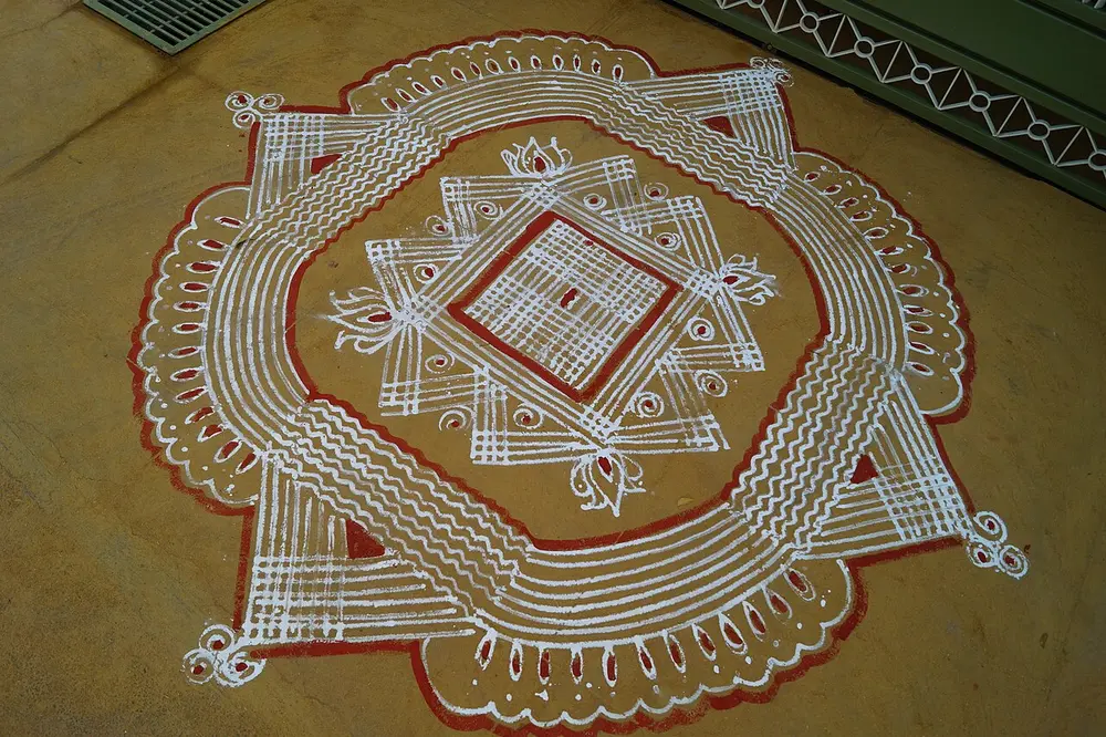 A large white line drawing on a swept floor, made of looping rice-flour lines that weave round a central square grid, with a thin red border painted round its outer edge.