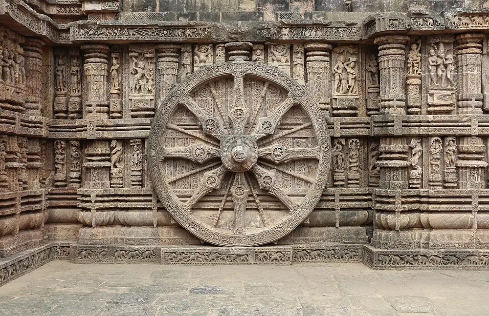 A huge carved stone chariot wheel set against the base of a temple wall, with eight thick spokes and eight thin ones round a big hub, and rows of small carved figures on the wall around it.