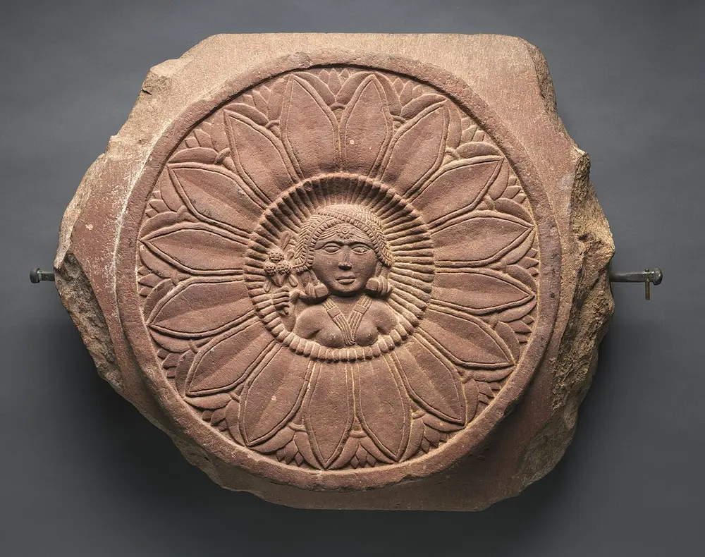 A round, reddish sandstone carving of a big open lotus with two rings of pointed petals, and the head and shoulders of a woman wearing a headdress at its centre.