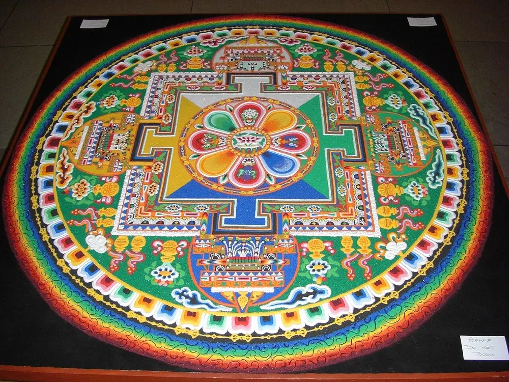 A large round design made of coloured sand on a table: inside rainbow-coloured outer rings sits a square palace with a gate in the middle of each side, and a red and blue eight-petalled lotus at its centre.