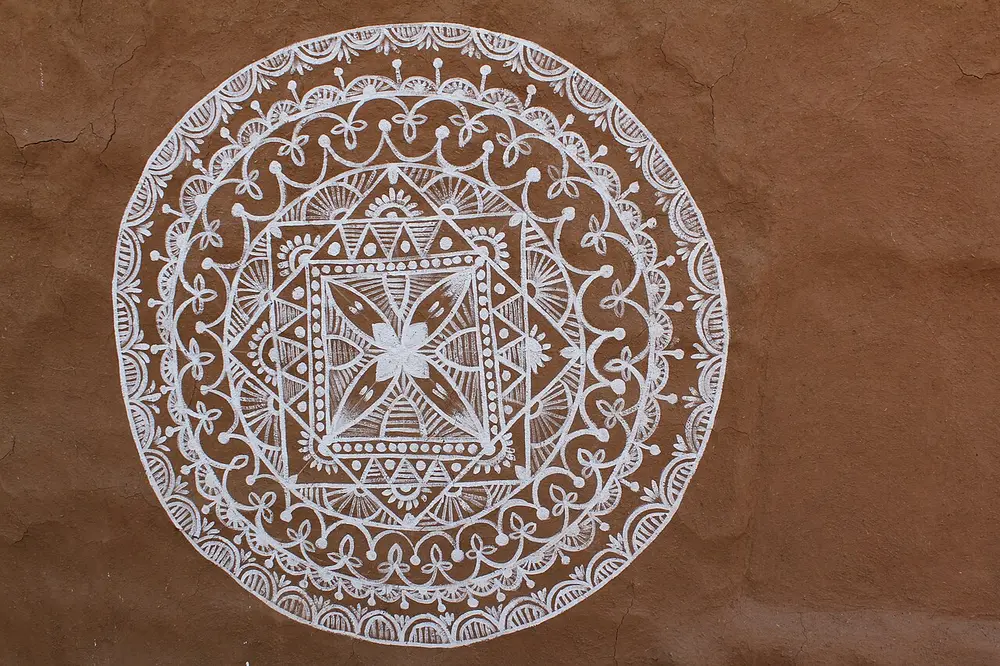 A round white design painted on a smooth brown mud wall, with a four-petalled flower inside a square, surrounded by rings of arches, dots, triangles and small loops.