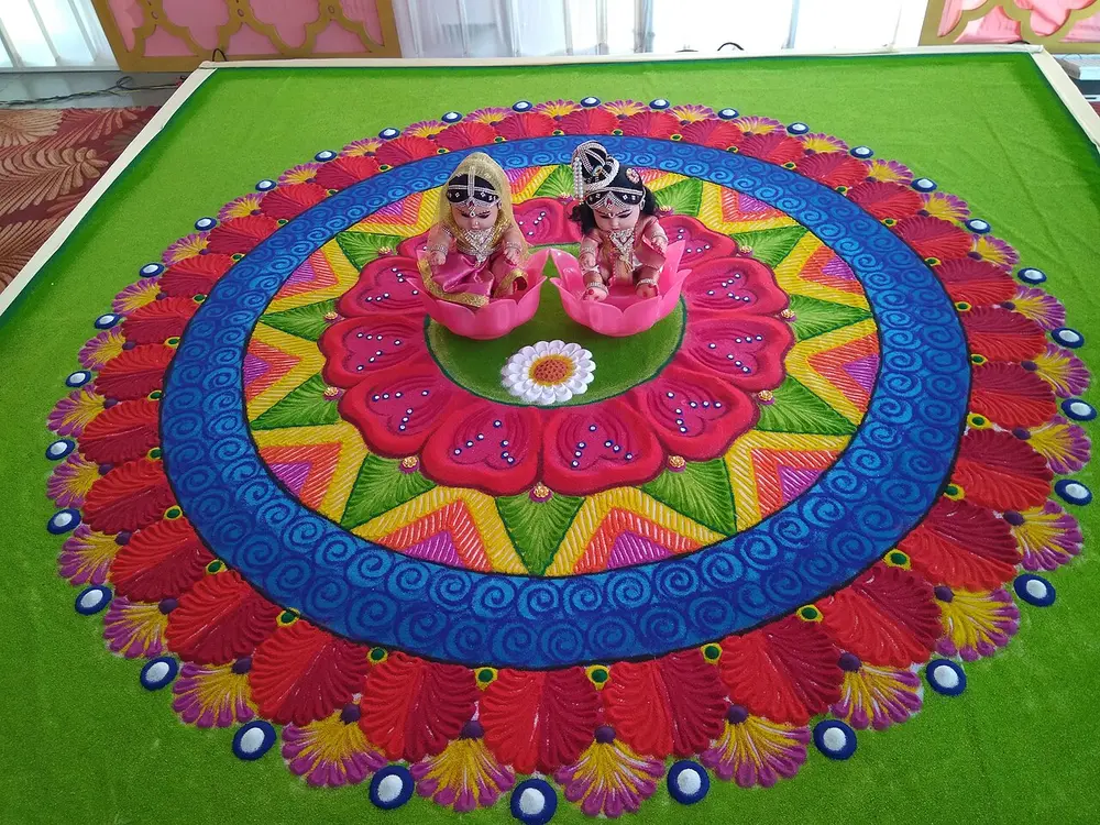 A large round floor design in bright coloured powders, with rings of red petals, blue and yellow zigzags and a pink lotus, and two small doll figures of a bride and groom at its centre, laid on a green mat.