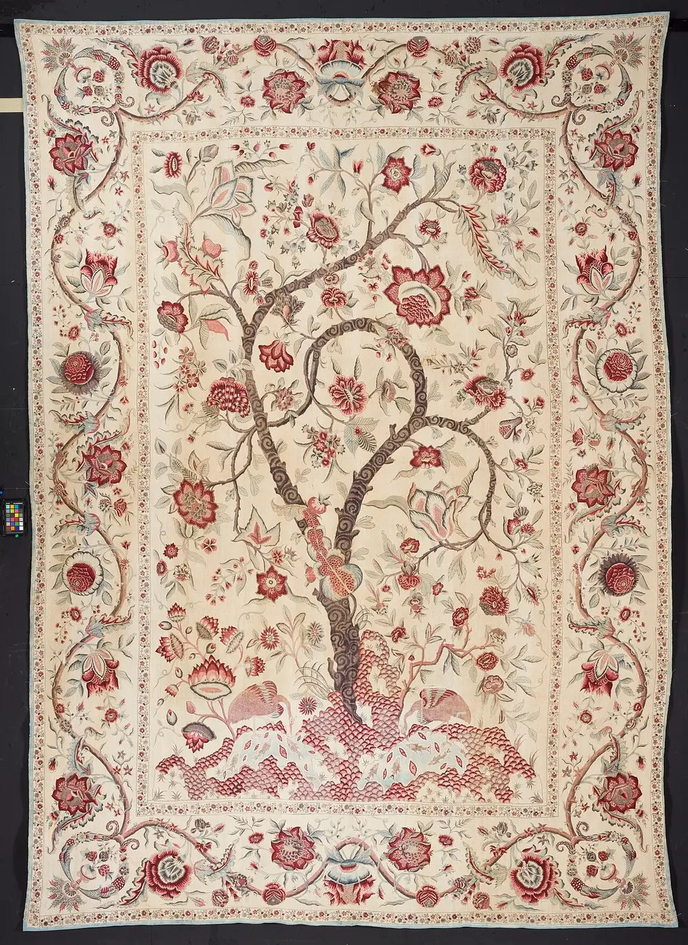 A large cream cotton bedcover painted in red, pink and brown with one tall, curving flowering tree growing from a small hill, birds and animals at its foot, and a wide border of flowering stems.