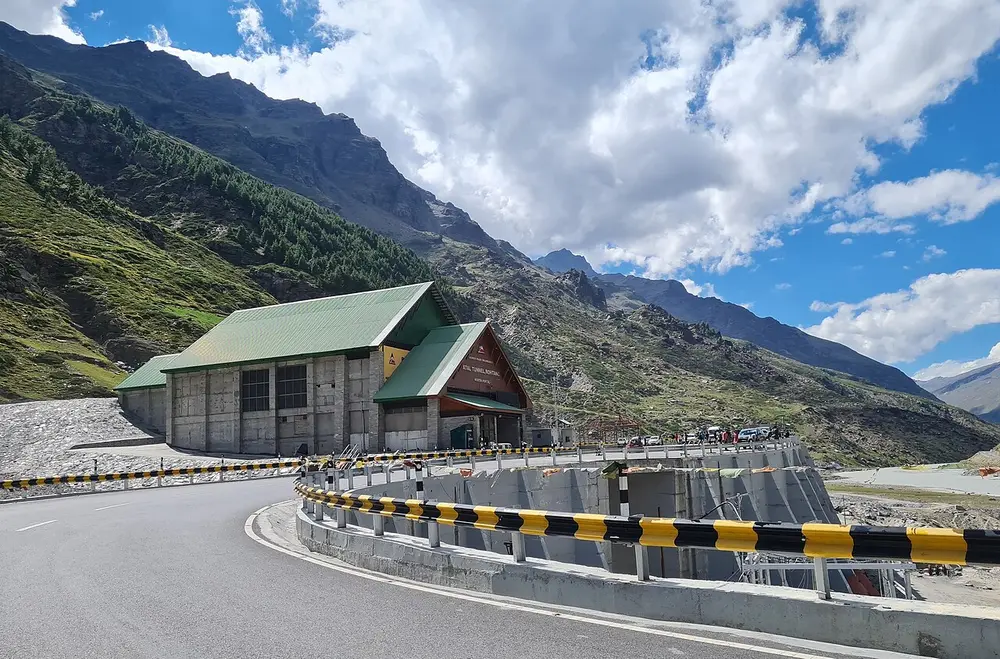 A road curving towards a tunnel entrance building at the foot of steep green mountains, under a blue sky with clouds.