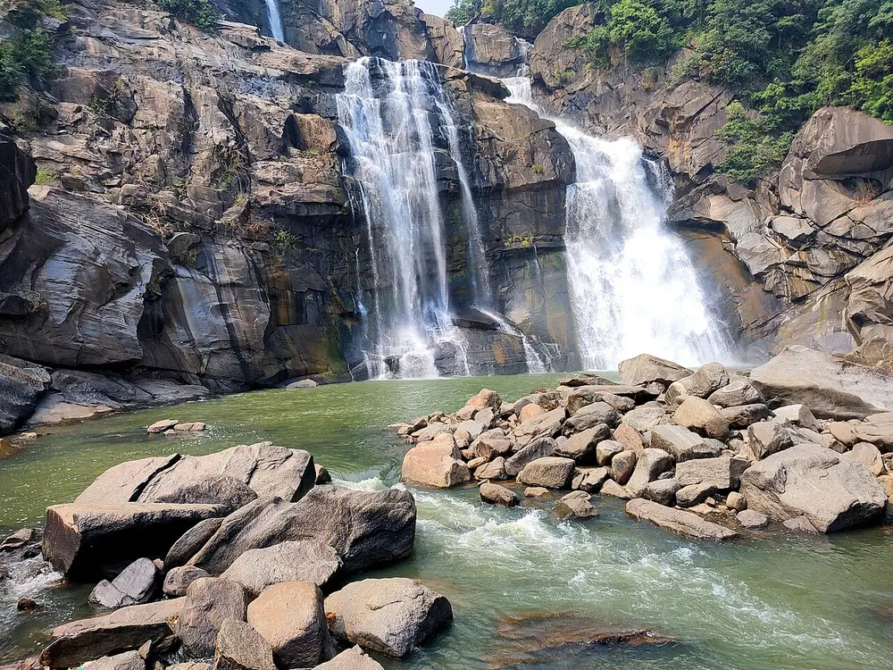 A waterfall pouring in several streams over dark layered rock into a green pool full of boulders.