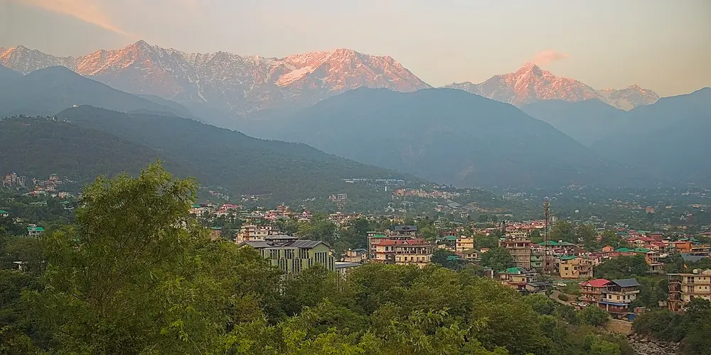 A row of snowy mountains glowing pink in the evening light, above a green hillside town.