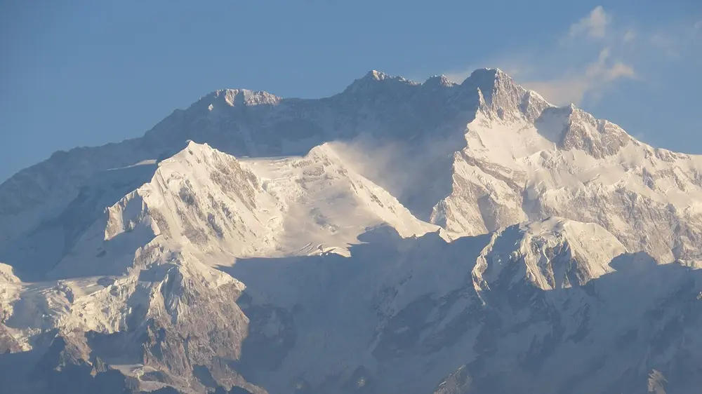 A massive snow-covered mountain wall with several summits, under a pale blue sky.