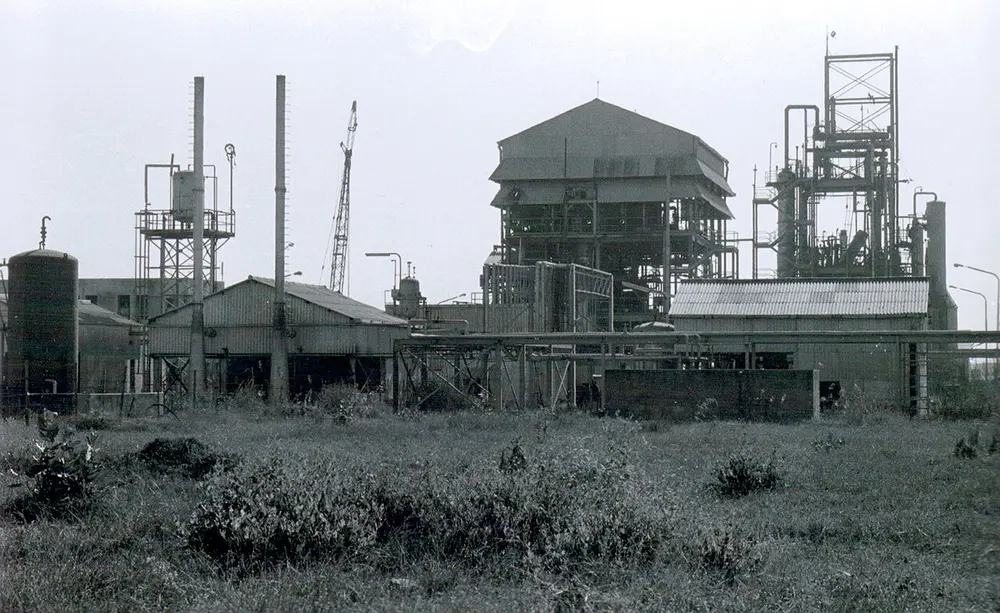 A black and white photo of an abandoned factory of tanks, pipes and metal towers, seen across a field of rough grass.