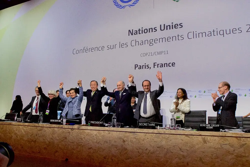 People on a stage raise their joined hands and cheer beneath a banner reading Conférence sur les Changements Climatiques 2015, Paris, France.