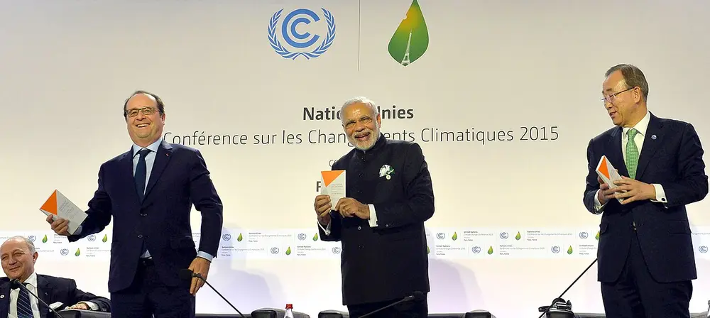 Three men in dark suits stand on a stage holding booklets, in front of a white backdrop with the UN climate logo and the words Conférence sur les Changements Climatiques 2015.
