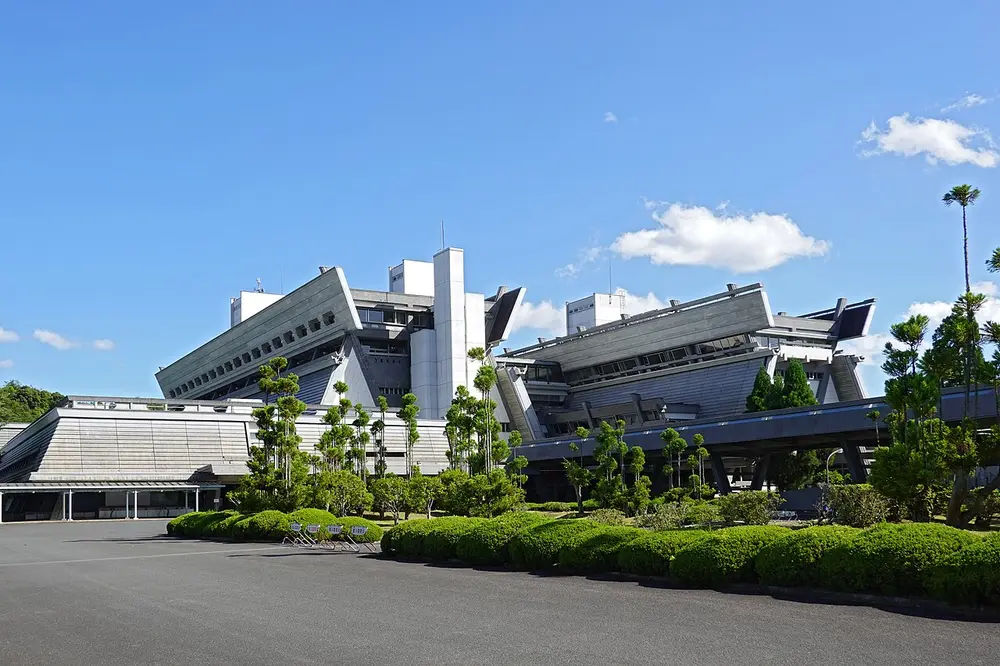 A large modern concrete building with steeply sloping walls and angled roofs, behind neat hedges, under a blue sky.