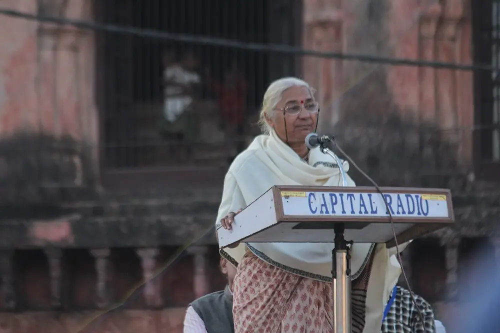 An elderly woman with white hair, wrapped in a white shawl, speaks into microphones at a lectern outdoors, in front of an old building.
