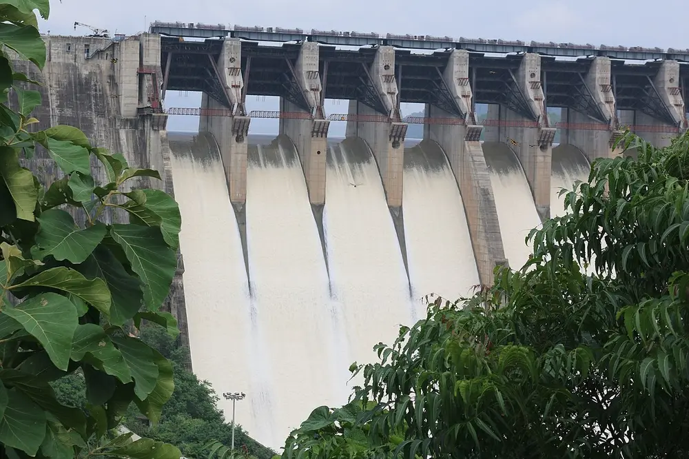 Water pours down the tall concrete spillway gates of a large dam, framed by green trees.