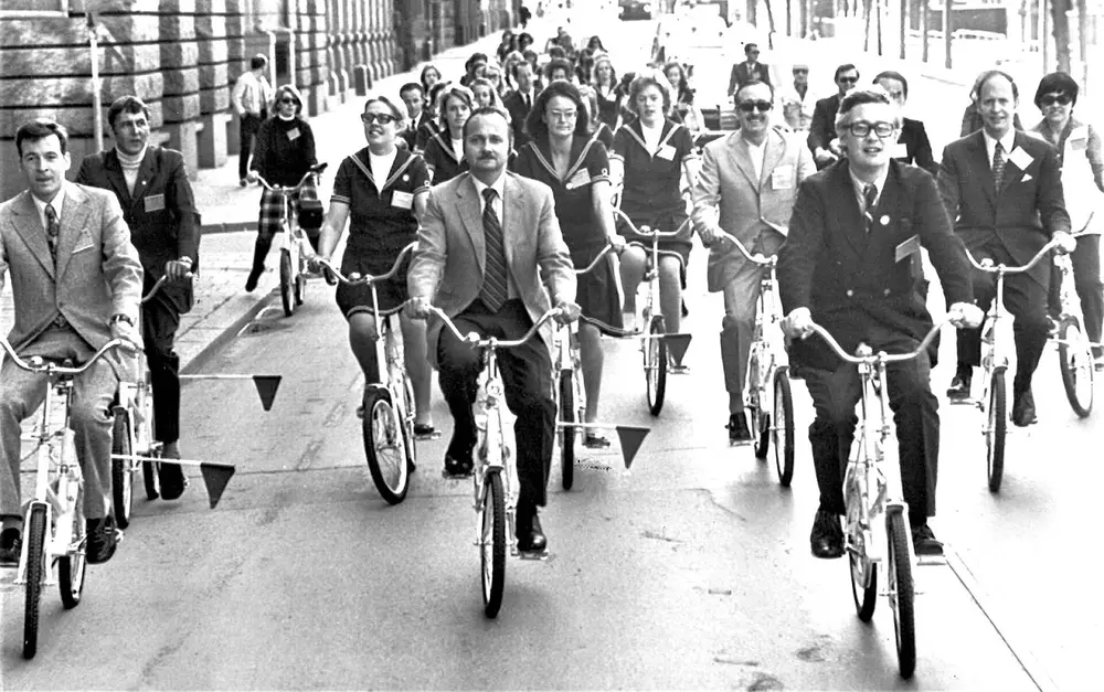 A black and white photo of men and women in suits riding bicycles together down a city street.