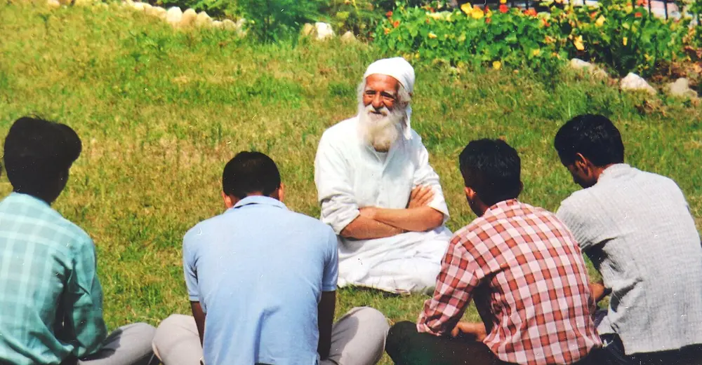 An old man with a long white beard, dressed in white, sits cross-legged on a lawn talking with four young men seated around him.