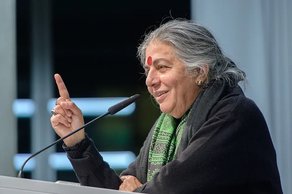 A woman with grey hair, a red bindi and a green scarf smiles and raises one finger as she speaks into a microphone.