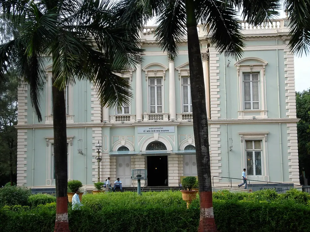 A pale green and white building with tall arched windows, columns and a balustrade along the roof, seen between palm trees across a garden.