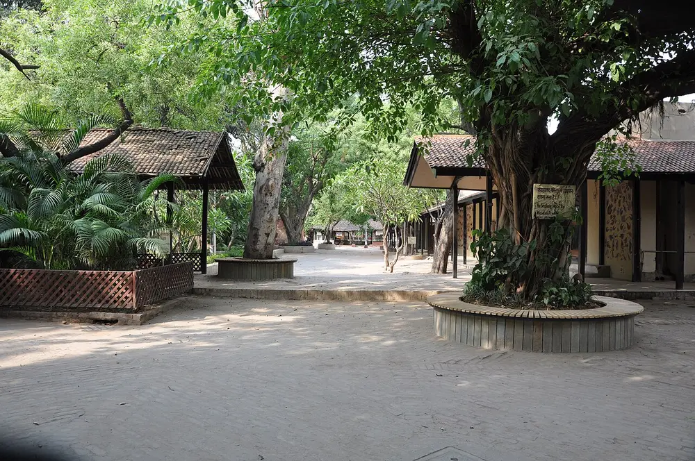 A shady paved courtyard with large old trees, a raised round planter and low buildings with sloping tiled roofs and verandahs.