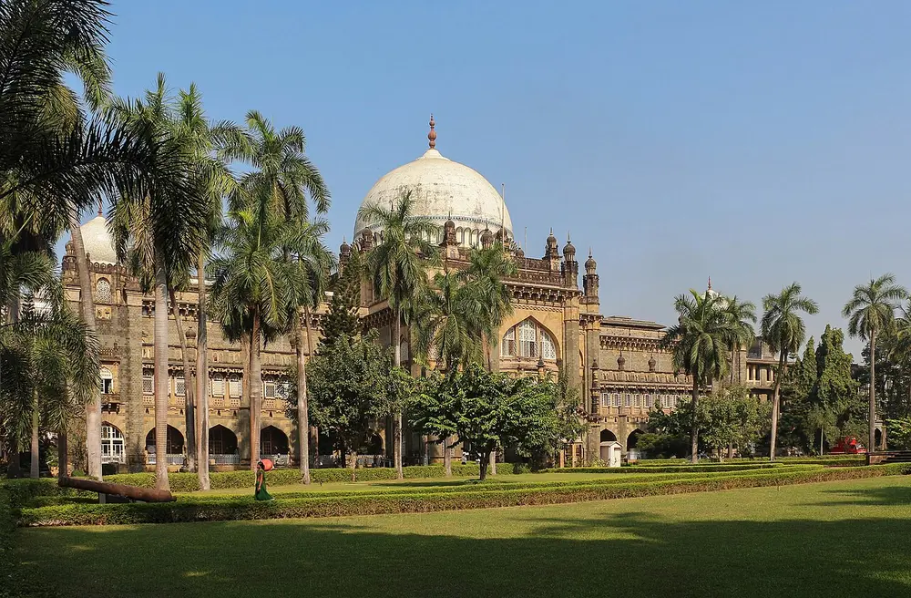 A grey stone building with a large white dome and smaller domed turrets, behind tall palm trees and a wide green lawn.