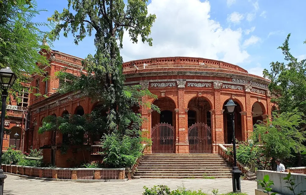 A round red-brick building with tall arched windows, cream trim and a wide flight of steps, half hidden by trees, in the museum complex at Egmore.