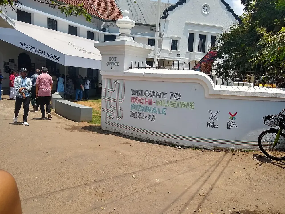 A white boundary wall painted with the words Welcome to Kochi-Muziris Biennale 2022-23, beside the gate of an old white bungalow where visitors queue under a canopy.