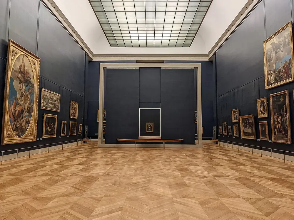A large, empty gallery with a glass ceiling and a wooden floor. Big framed paintings hang on dark blue walls, and the small Mona Lisa hangs alone in the middle of the far wall.