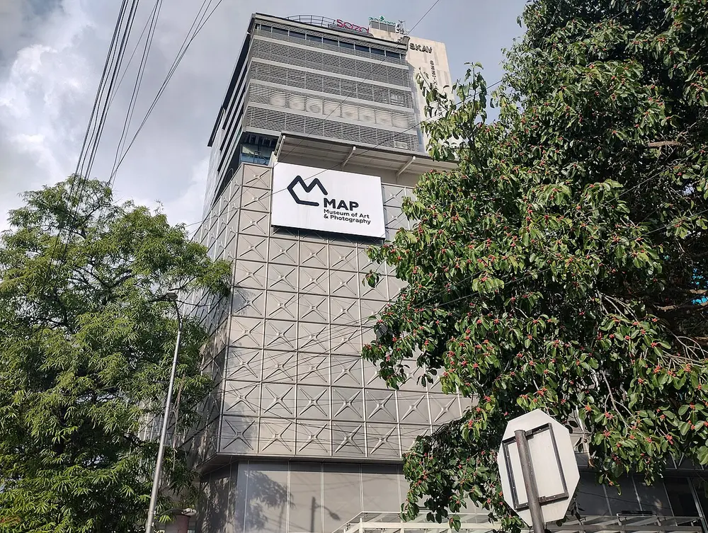 A tall grey building covered in a lattice of square panels, with a white sign showing the MAP logo, partly framed by green trees.