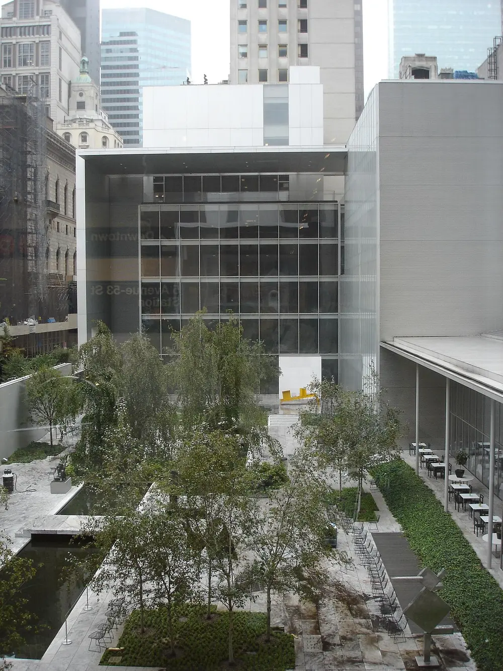 Looking down into a garden of slim trees and paving between tall glass and grey stone museum buildings, with skyscrapers behind.