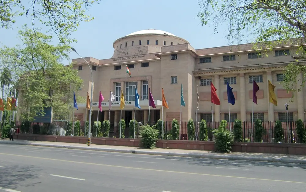 A long, low sandstone-coloured building with a round drum-shaped tower at the corner and a row of colourful flags in front, beside a wide road.