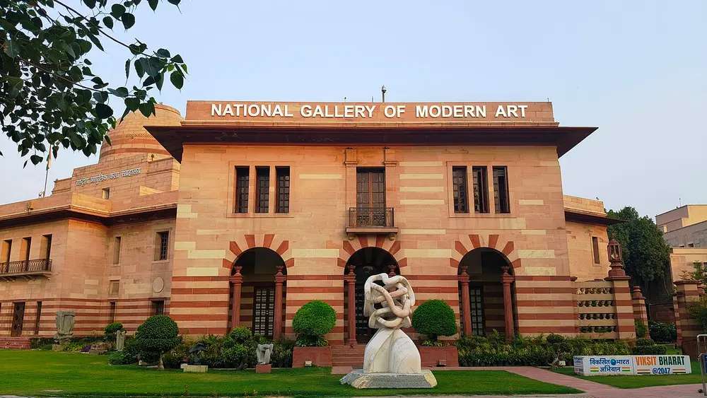 A red and cream sandstone building with a low dome, arched windows and the words National Gallery of Modern Art across the top, with a white abstract sculpture on the lawn in front.