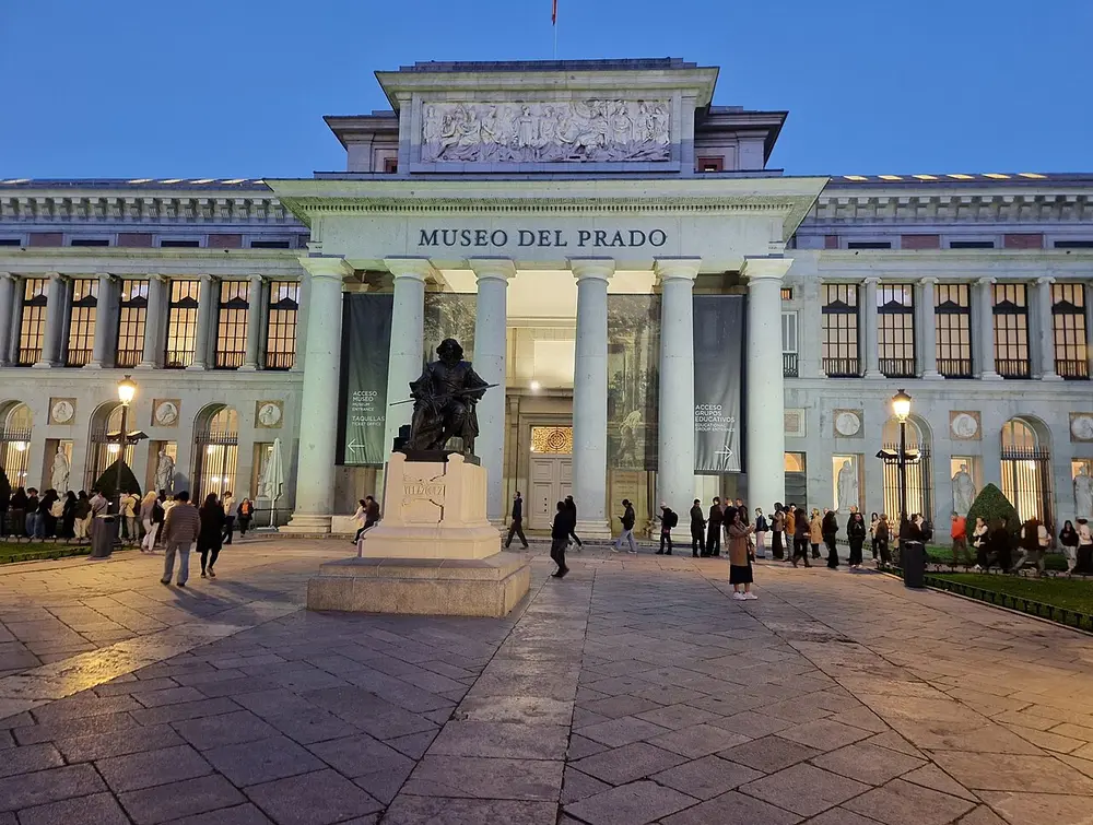 The columned entrance of a long stone museum with the words MUSEO DEL PRADO above the door, a bronze statue of a seated man on a tall base in front, and visitors in the square at dusk.