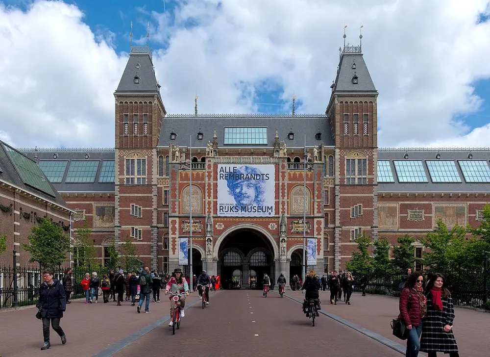 A large red-brick building with two pointed towers, many windows and a tall arched passage in the middle, with people walking and cycling towards it.