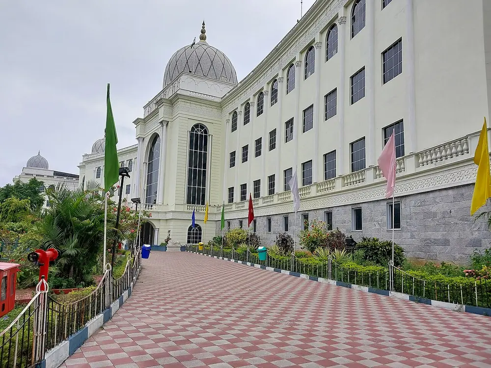 A long white museum building with rows of windows and a large white dome at one end, approached by a paved path lined with flags and plants.
