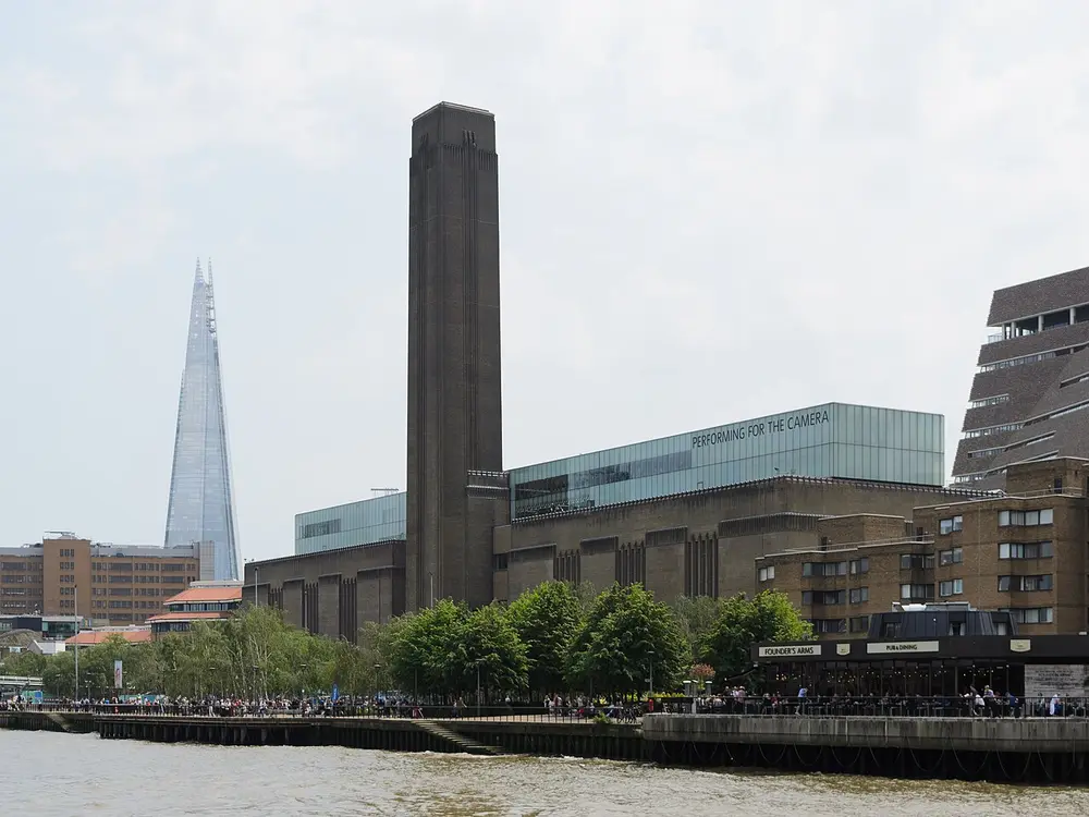 A long brown-brick former power station with one tall square chimney in the middle and a modern brick extension beside it, seen across the River Thames, with the pointed Shard skyscraper behind.
