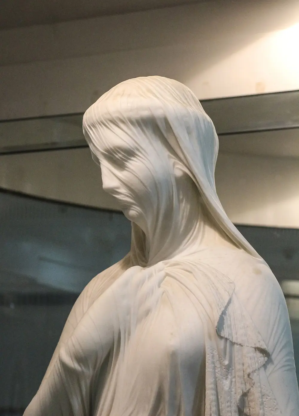 A close view of a white marble statue of a young woman whose head and face are covered by a thin veil, carved so finely that her features show through it.
